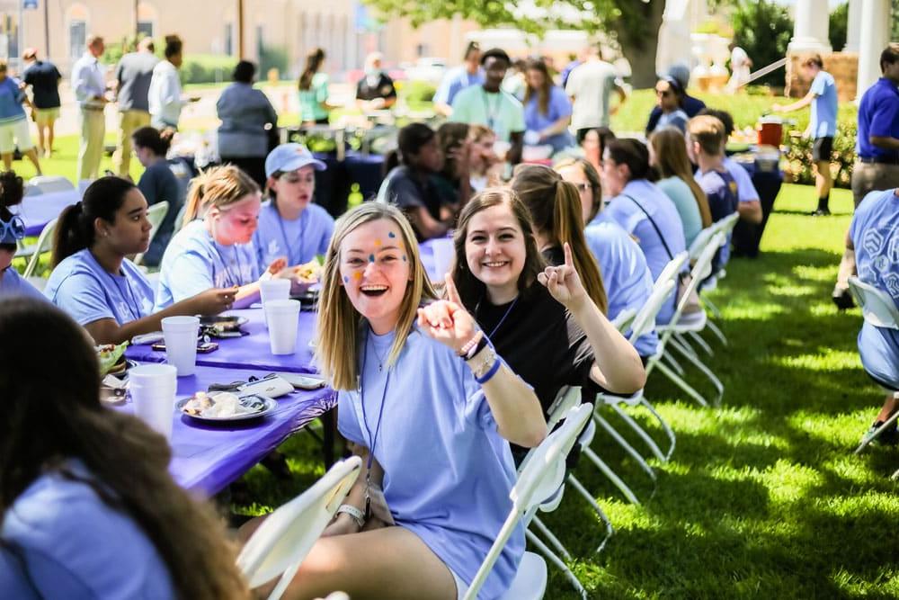 Group of Howard Payne University people wearing blue shirts enjoying an outdoor gathering with food and drinks on a sunny day. | HPU